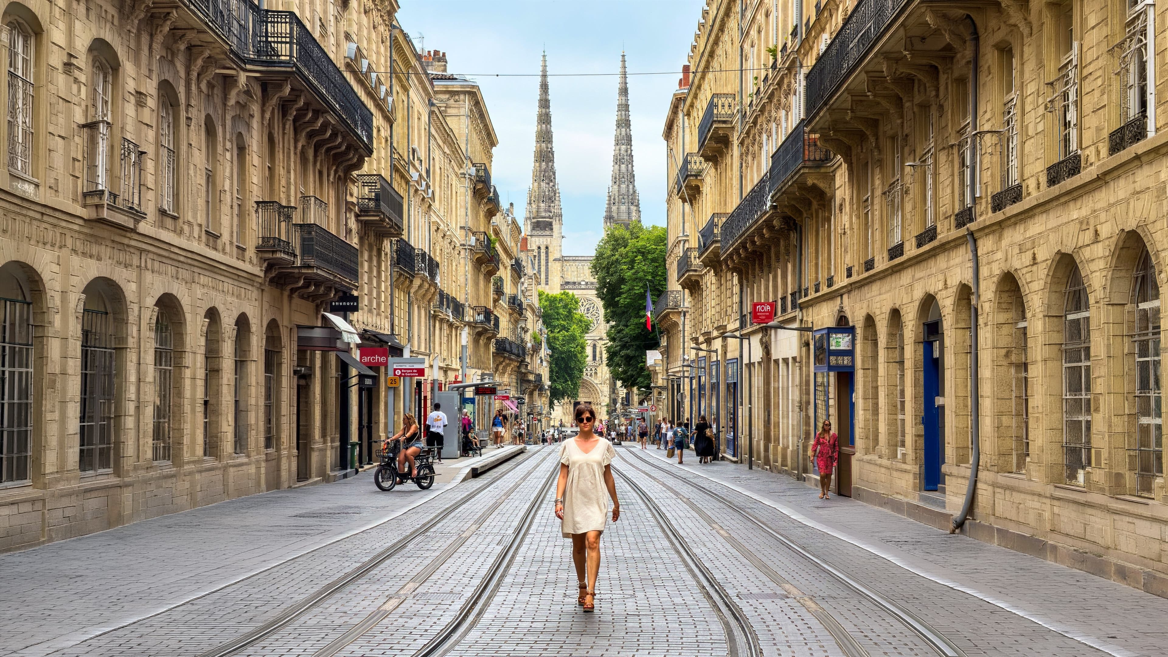Femme en robe courte beige marchant dans une rue pavée de Bordeaux, flèches de la cathédrale Saint-André en perspective.
