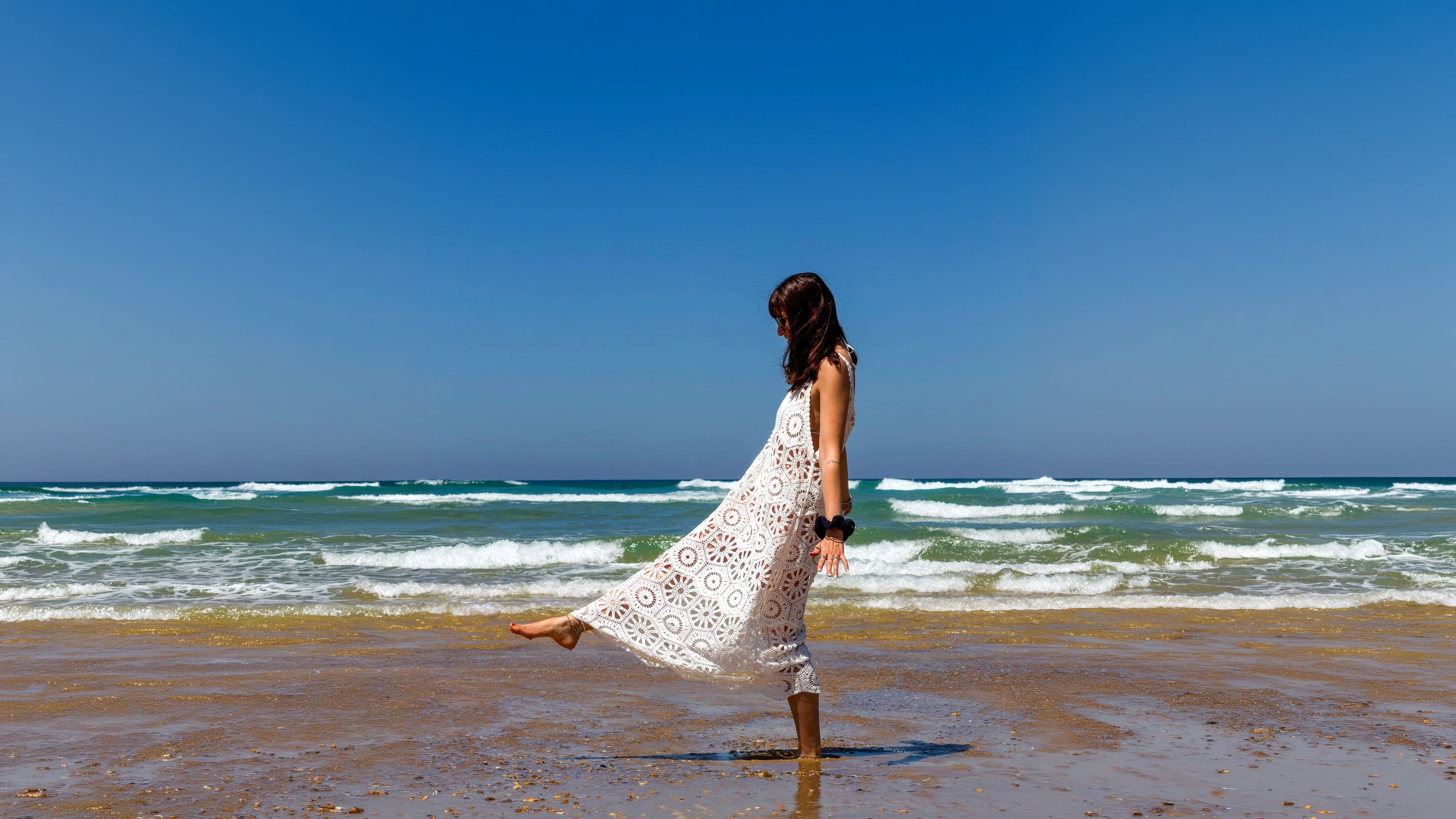 Femme en robe longue blanche en crochet ajouré au bord de l'océan, vagues et ciel bleu à l'horizon.