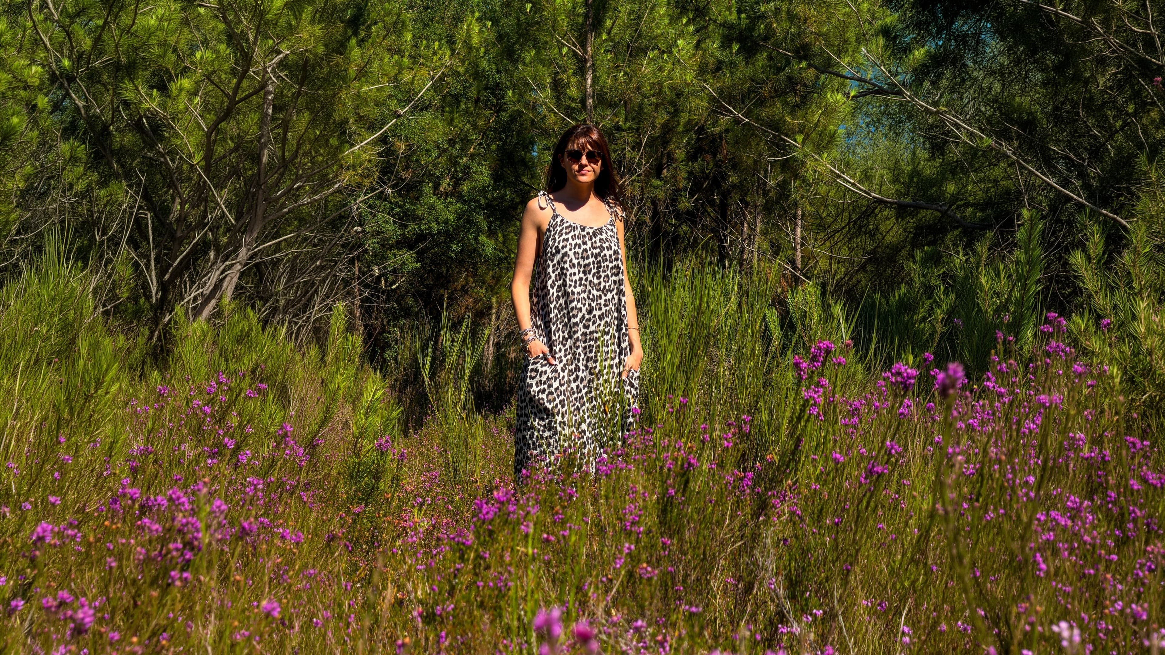 Femme en robe longue à imprimé léopard, debout dans un champ de bruyère mauve en fleurs, forêt de pins en arrière-plan.