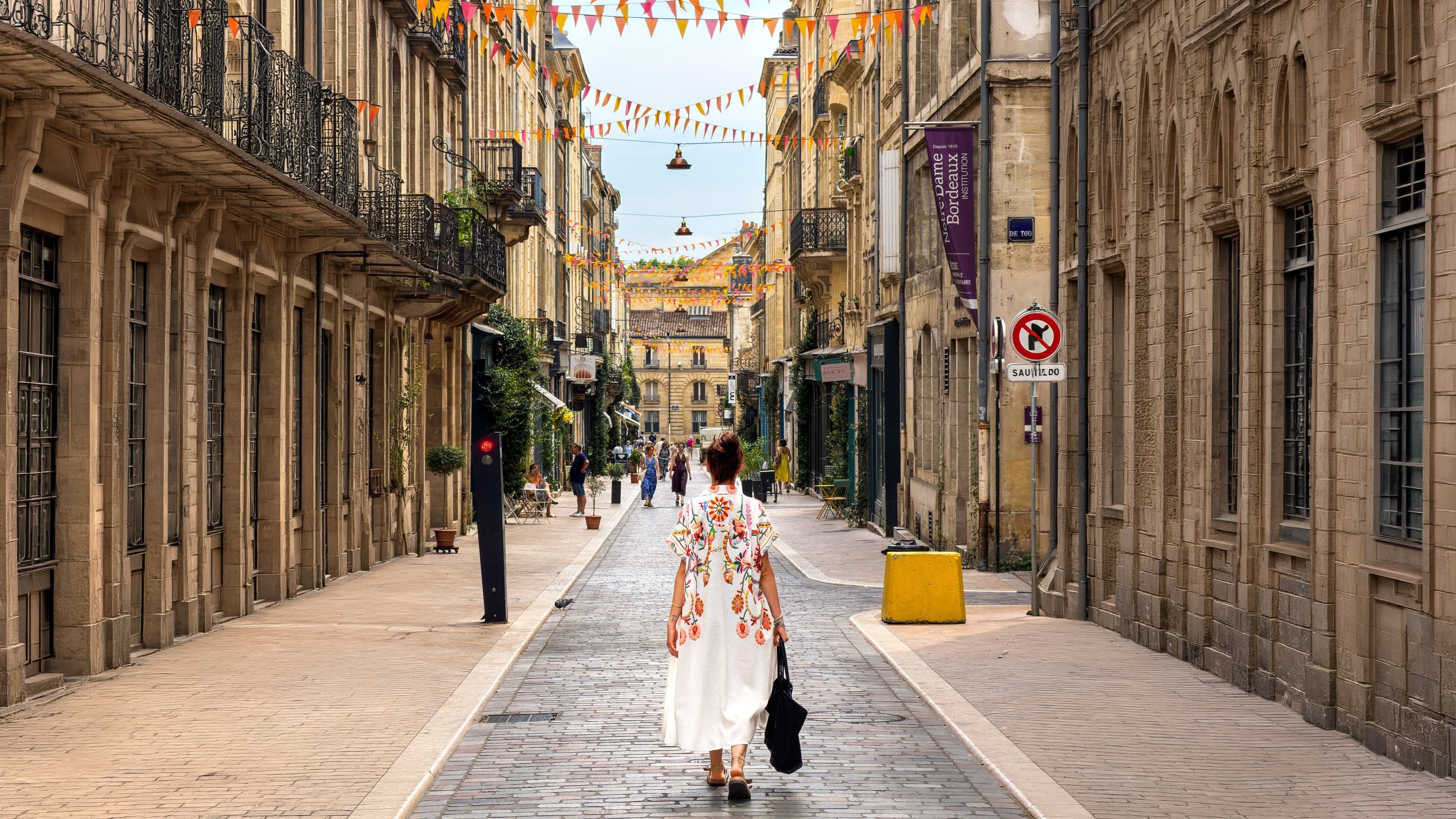 Femme de dos en robe longue blanche à motifs floraux, sac à la main, dans une rue de Bordeaux décorée de guirlandes de fanions colorés.